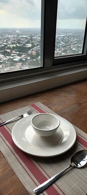 A dining setup with a plate, bowl, fork, and spoon on a mat, with a window view of a cityscape. - Cozy High-Rise Haven (Quezon City)