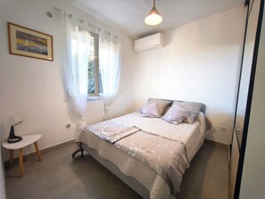 Ein Schlafzimmer mit einem Bett, einem Nachttisch, einer Lampe, einem Fenster mit Vorhängen und einer Klimaanlage.