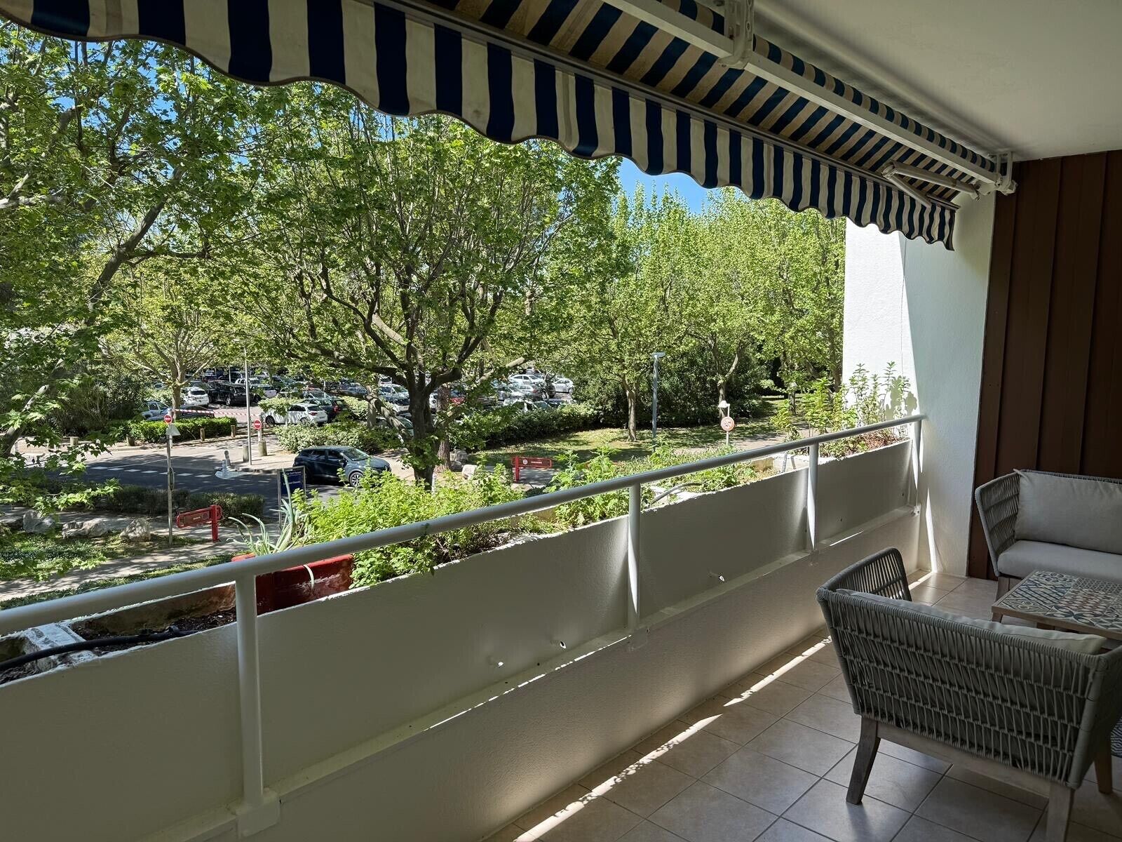 Un balcon donnant sur des arbres et des voitures garées, avec une balustrade blanche et un fauteuil en osier.