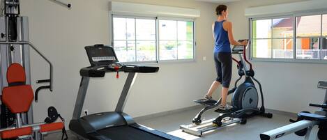 Un gimnasio con equipo de ejercicio, ventanas amplias y una persona usando una cinta de correr.