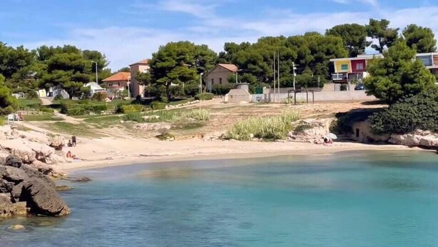 Uma área costeira com águas cristalinas, praia de areia e edifícios ao fundo.