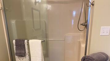 A bathroom with a glass shower enclosure, a toilet, a sink, and a small wooden shelf with towels and toilet paper.