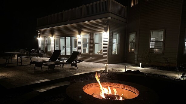 A house with a fire pit in the foreground, outdoor seating, and a well-lit patio area at night.