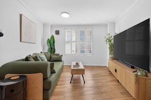 Ein Wohnzimmer mit einem grünen Sofa, einem Couchtisch, einem Fernseher auf einer Holzkommode und einem Fenster mit Jalousien.