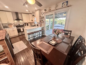 A modern kitchen with a central island, bar stools, a dining table with chairs, and a view of the outdoors through a glass door.