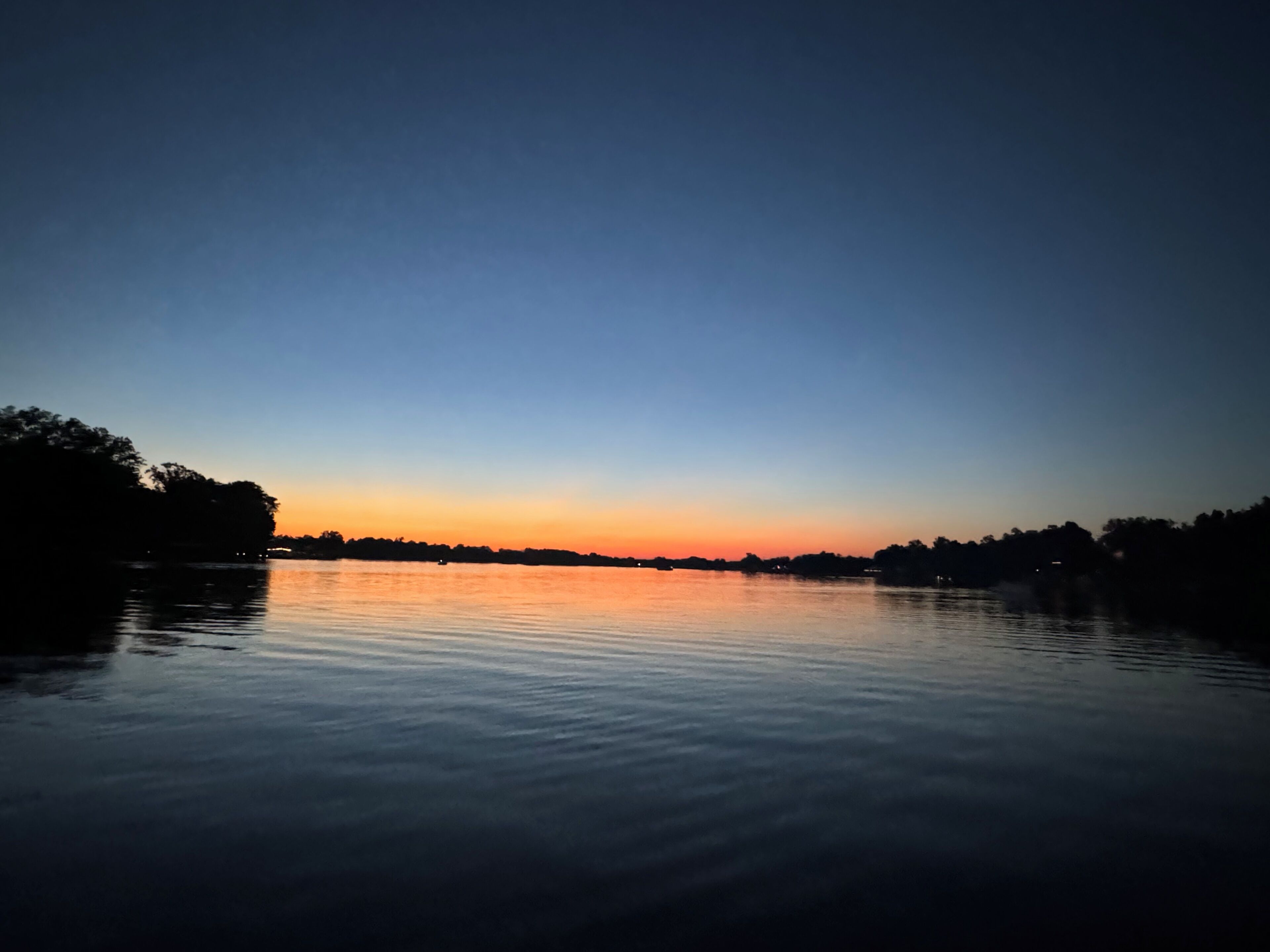 A calm lake at dusk with a clear horizon and trees silhouetted against the sky.