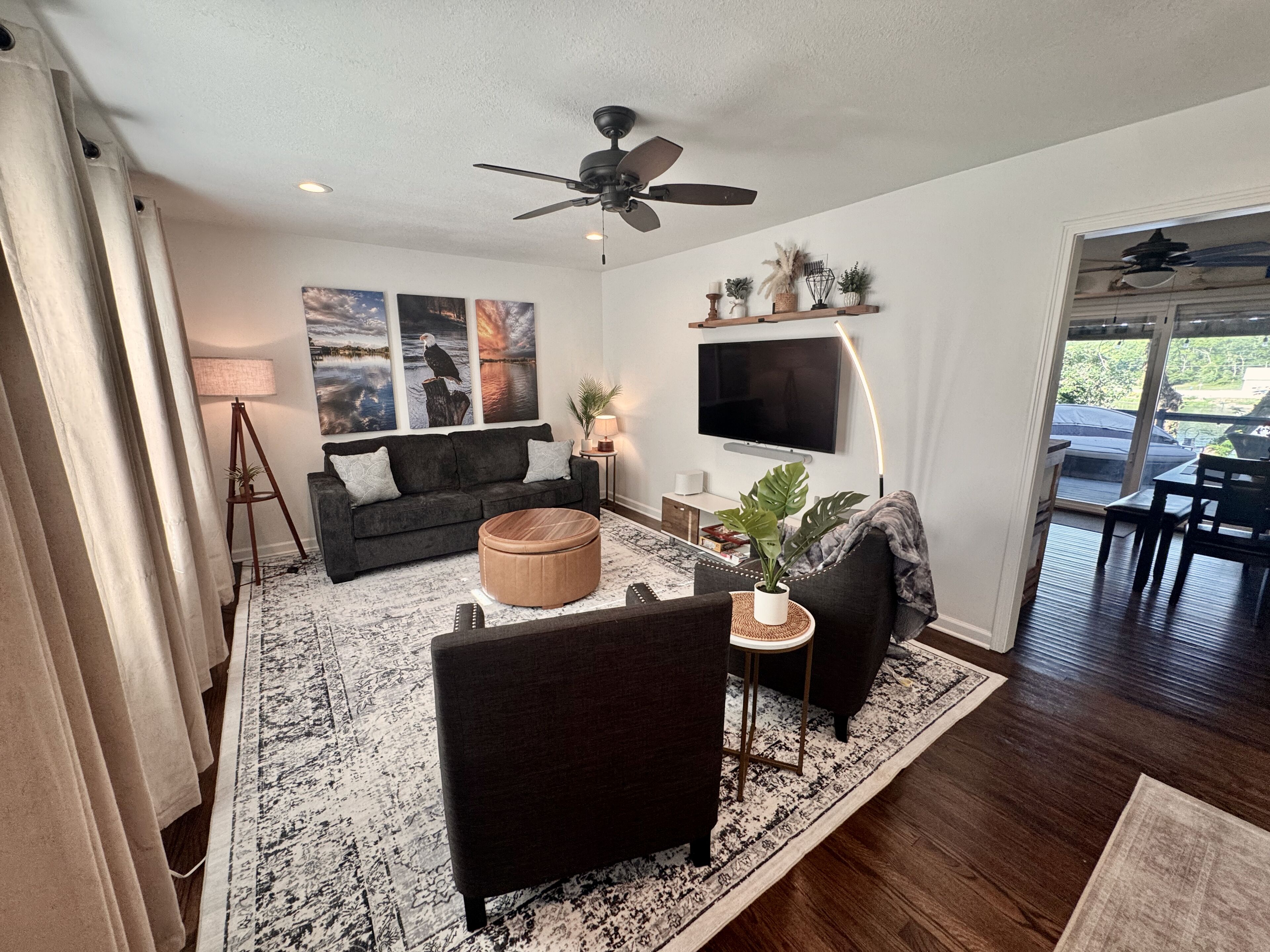Un salon moderne avec un canapé foncé, un pouf rond et une table basse. Un téléviseur à écran plat est fixé au mur, ainsi qu’un ventilateur de plafond. Une grande fenêtre offre une vue sur l’extérieur.
