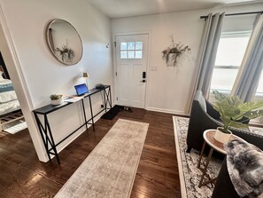 A well-lit hallway with a desk, a laptop, and a door. There are two hanging plants and a potted plant on a side table.