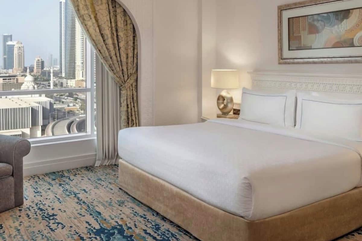 A hotel room with a large bed, a city view, a painting on the wall, and a patterned carpet.