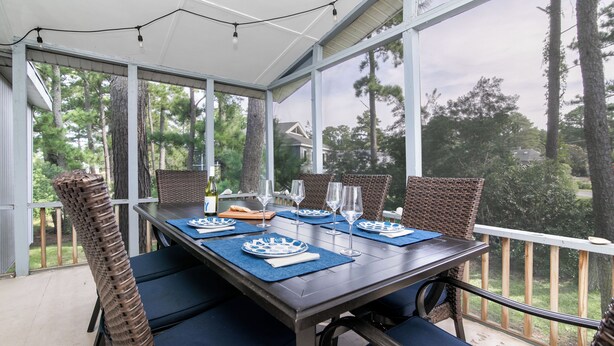 A covered outdoor dining area with a table set for four, surrounded by wicker chairs, and overlooking a wooded area. - Pearl in the Pines – Bikes, Kayaks, EV Charger, Screened Porch, Fire Pit, Pets (Chincoteague)