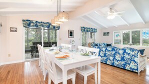 A spacious room with a dining table, chairs, a sofa, and a large window. - Pearl in the Pines – Bikes, Kayaks, EV Charger, Screened Porch, Fire Pit, Pets (Chincoteague)