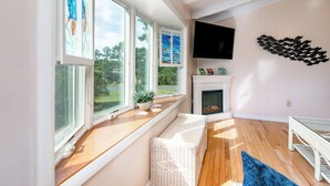 A room with large windows, a fireplace, a television, and a white wicker bench. - Pearl in the Pines – Bikes, Kayaks, EV Charger, Screened Porch, Fire Pit, Pets (Chincoteague)