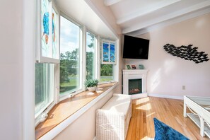 Ein Zimmer mit großen Fenstern, einem Kamin, einem Fernseher und einer weißen Rattanbank.