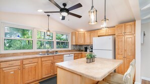 A modern kitchen with wooden cabinets, a central island, a ceiling fan, and large windows. - Pearl in the Pines – Bikes, Kayaks, EV Charger, Screened Porch, Fire Pit, Pets (Chincoteague)