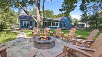 A backyard with a stone fire pit, wooden Adirondack chairs, and a blue house.