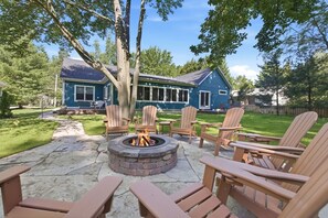 Ein Garten mit einem Steinkamin, Holz-Adirondack-Stühlen und einem blauen Haus.