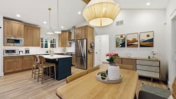 A modern kitchen with wooden cabinets, stainless steel appliances, a dining area with a wooden table, and pendant lighting.