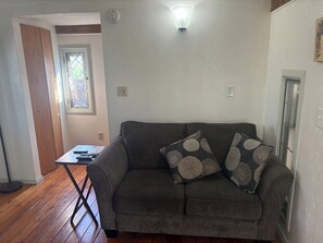 Ein Wohnzimmer mit grauem Sofa, gemusterten Kissen, einem kleinen Tisch mit Fernbedienungen, Holzboden und einem Fenster mit Blick.