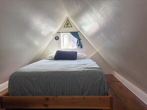 Ein Einzelbett in einem Raum mit schrägen Decken, einem dreieckigen Warnschild, einem Fenster mit Vorhängen und einem Holzboden.