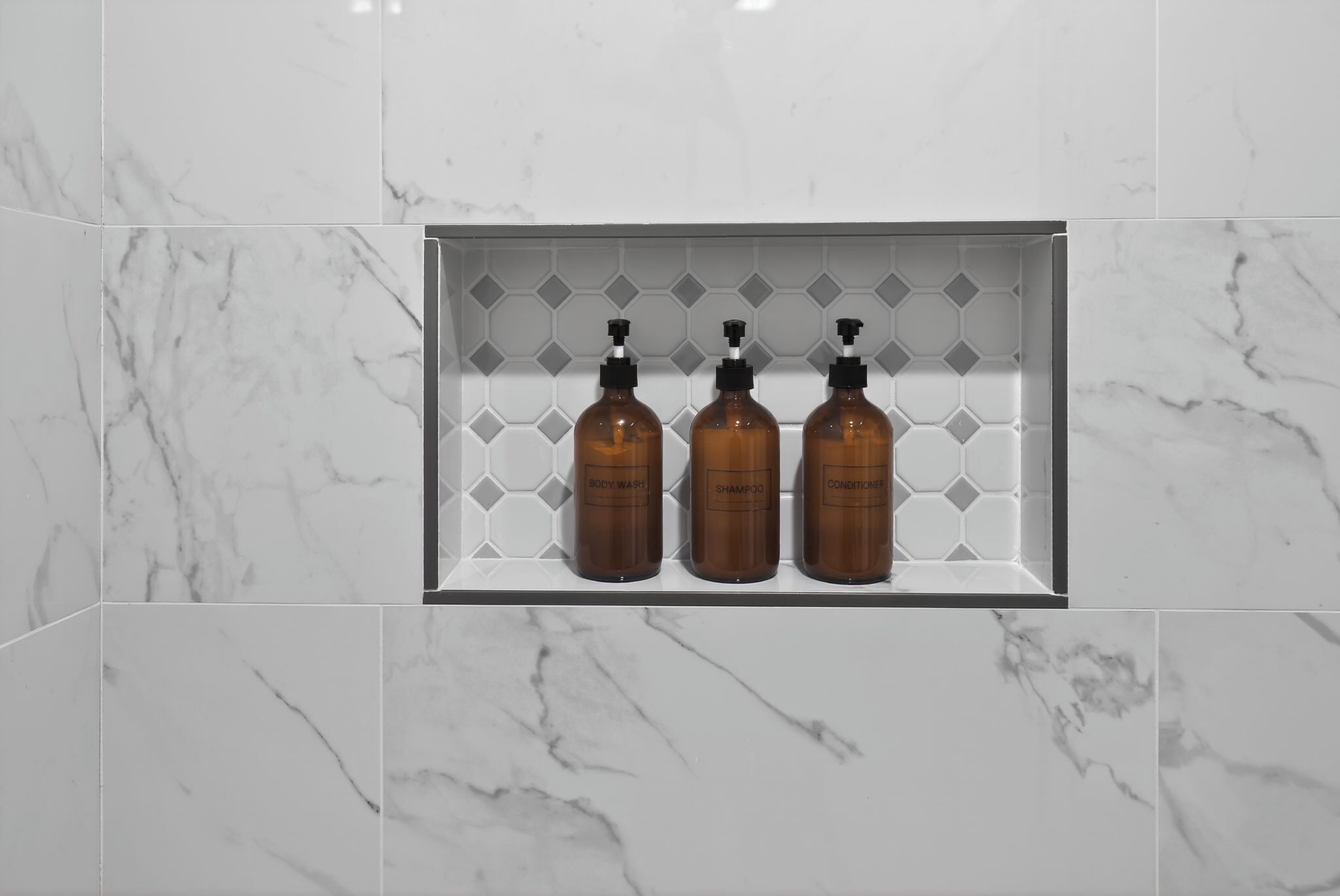 A bathroom with three brown bottles labeled body wash, shampoo, and conditioner in a white tiled sink area.