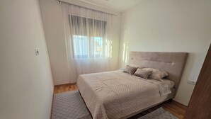 A bedroom with a bed, a window with curtains, a wooden floor, and a door. - Urban Comfort Apartments, Podgorica (Podgorica)