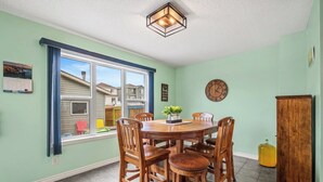 A dining room with a round wooden table, six chairs, a clock, and a window with a view of neighboring houses.