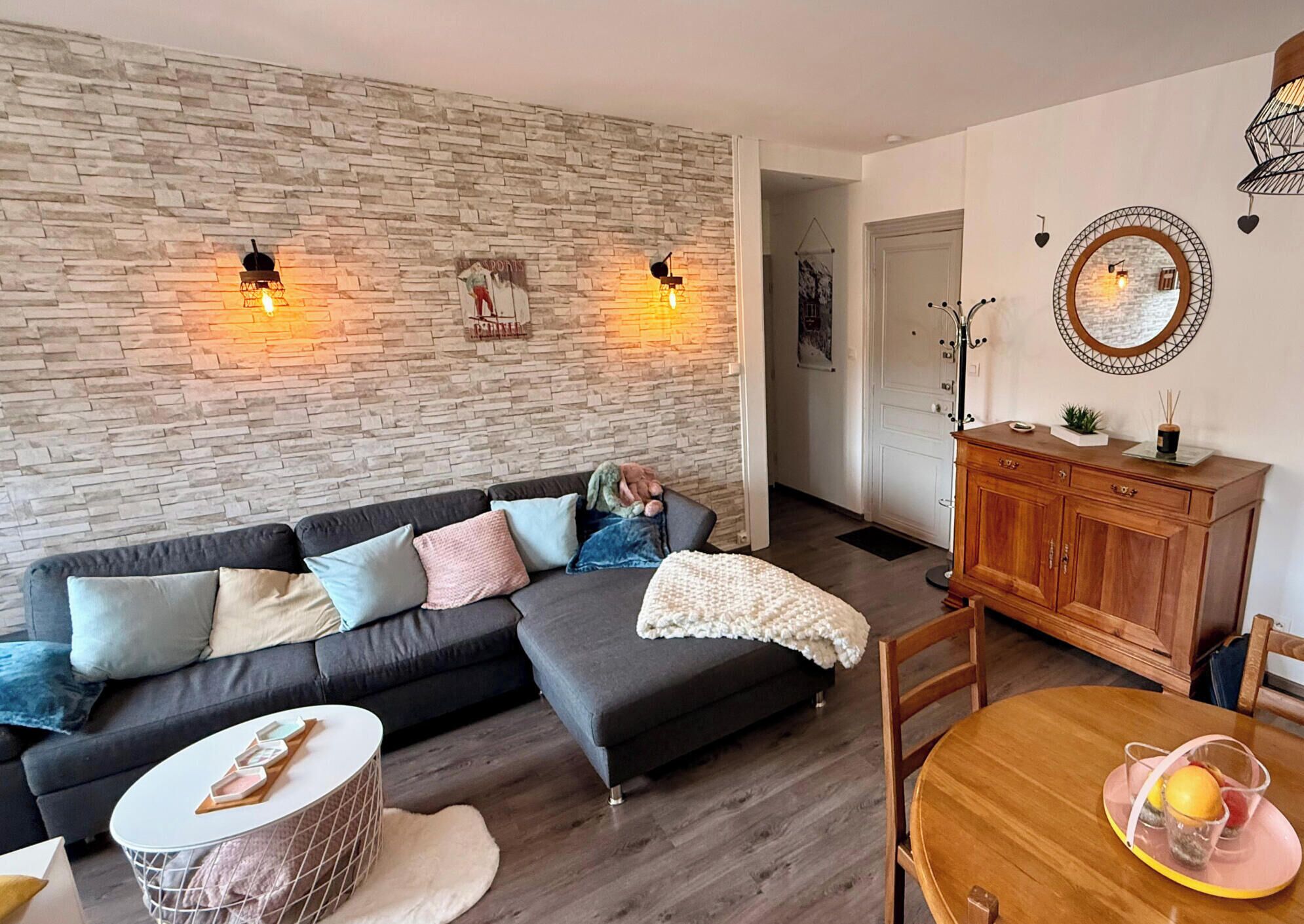 Un salon avec un mur en pierre, un canapé gris foncé, un meuble en bois et une table à manger agrémentée d’un plateau rose.