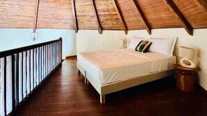 Quarto com teto inclinado, vigas de madeira, uma cama, mesa de cabeceira e corrimão junto à janela.