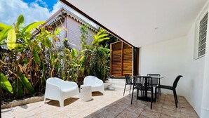 Um pátio externo com cadeiras e mesa de plástico branco, cercado por plantas tropicais e um muro de madeira.
