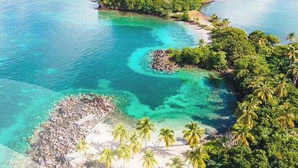 Uma ilha tropical com águas turquesa cristalinas, vegetação exuberante e uma pequena área de praia rochosa.