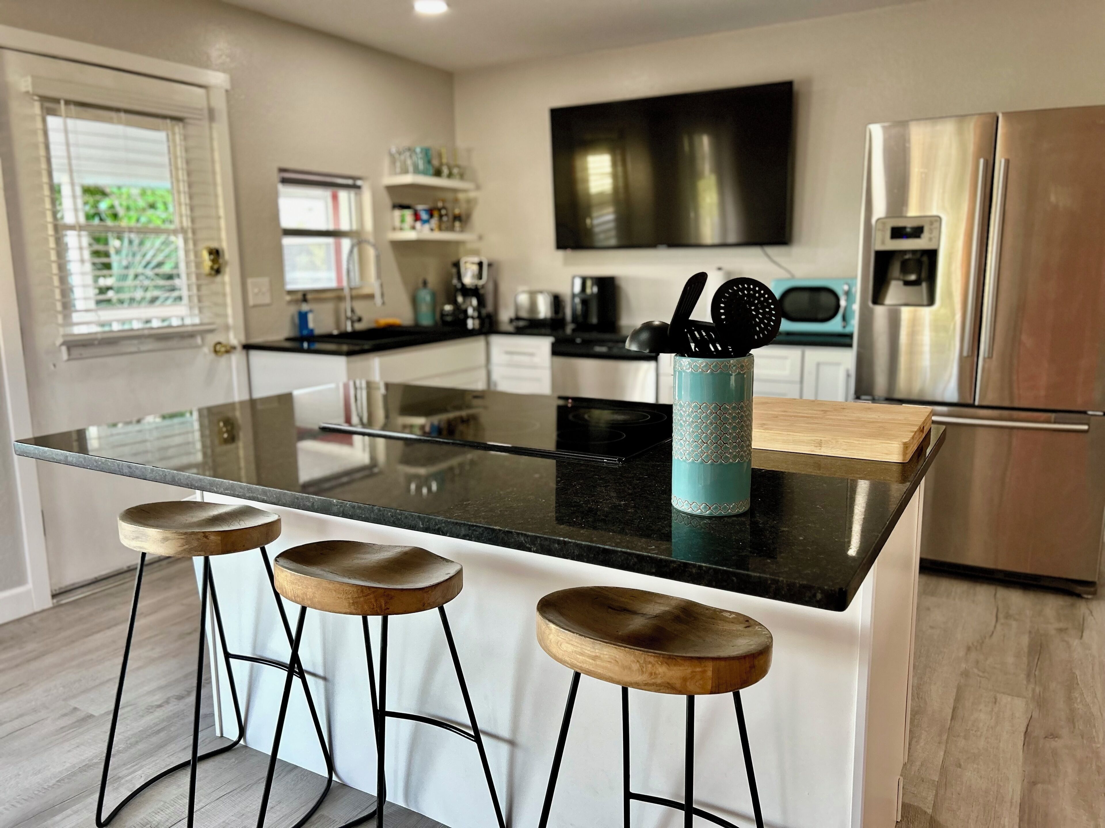 A modern kitchen with a glass island, wooden stools, a stainless steel refrigerator, and a flat-screen TV.