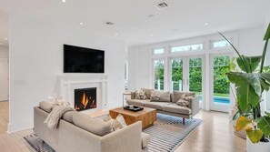A modern living room with a fireplace, a flat-screen TV, a sofa, a coffee table, and a large window with a view of greenery. - Exquisite Shinnecock Hills Classic, Private Yard featuring Heated Pool,... (Southampton)
