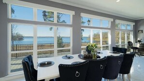 A dining area with a table and chairs, large windows with a view of a body of water, and a balcony with a railing. - Lakefront Estate Sleeps 18, Bunk & Game Room & Boat House w/Swim Hole & Kitchen! (Pittsburg)