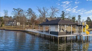 A waterfront house with a dock extending into the water, a covered patio, and a yellow slide. - Lakefront Estate Sleeps 18, Bunk & Game Room & Boat House w/Swim Hole & Kitchen! (Pittsburg)
