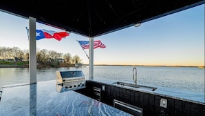 A covered outdoor area with a grill and sink, flags, and a view of a body of water. - Lakefront Estate Sleeps 18, Bunk & Game Room & Boat House w/Swim Hole & Kitchen! (Pittsburg)
