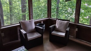 Uma cabana de madeira com duas cadeiras de vime e uma pequena mesa, janelas amplas com vista para a floresta.