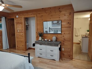 A wooden-paneled room with a bed, a white dresser, a ceiling fan, a painting, and a bathroom visible through an open door.