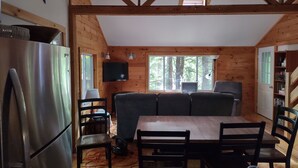 Interior de uma cabana de madeira com uma mesa de jantar, cadeiras, um sofá, uma televisão e um frigorífico.
