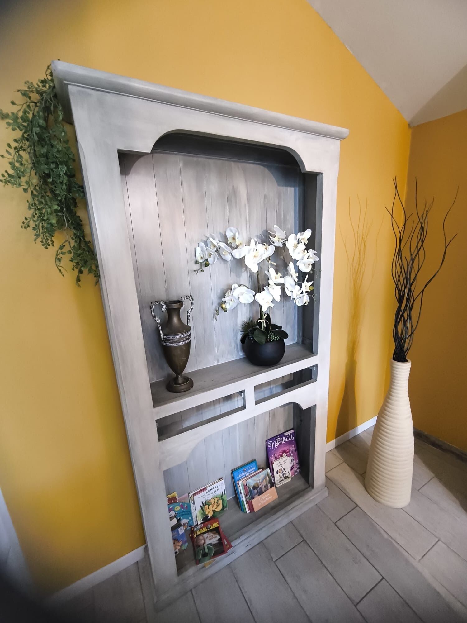 Une cheminée décorée de fleurs blanches, un vase et des étagères à livres, le tout adossé à un mur jaune.