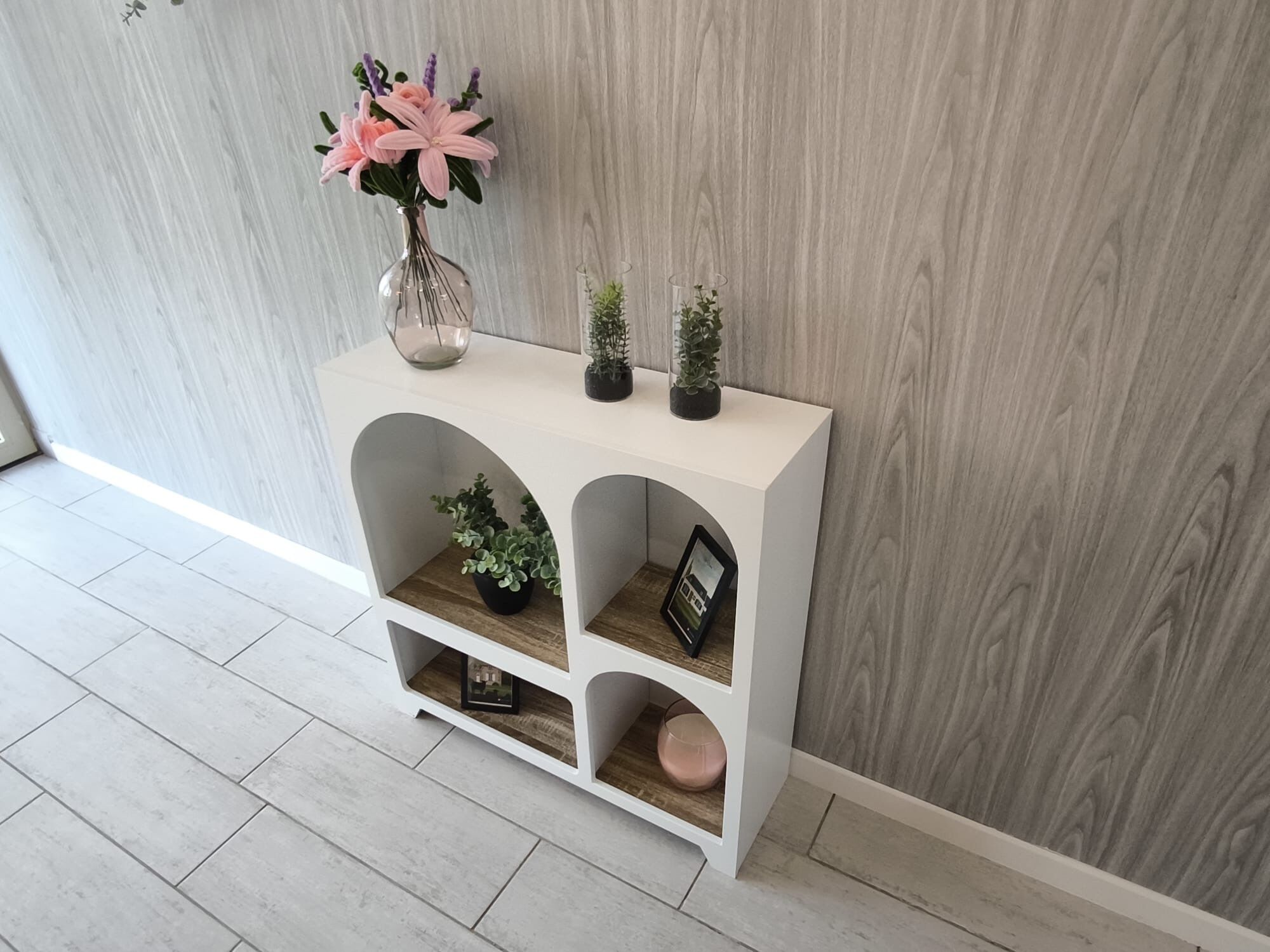 A white console table with three shelves, each containing a plant and a small item.