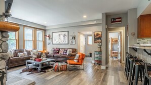 Een ruime woonkamer met een accentmuur van steen, een groot raam, een open haard en een keuken met barstoelen.