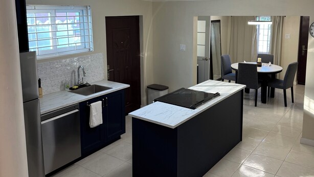 A modern kitchen with a central island, a dining area with a table and chairs, and a view of the outside through a window. - Stay Exchange Modern Chill Retreat w/ Private Backyard Oasis (Ocho Rios)