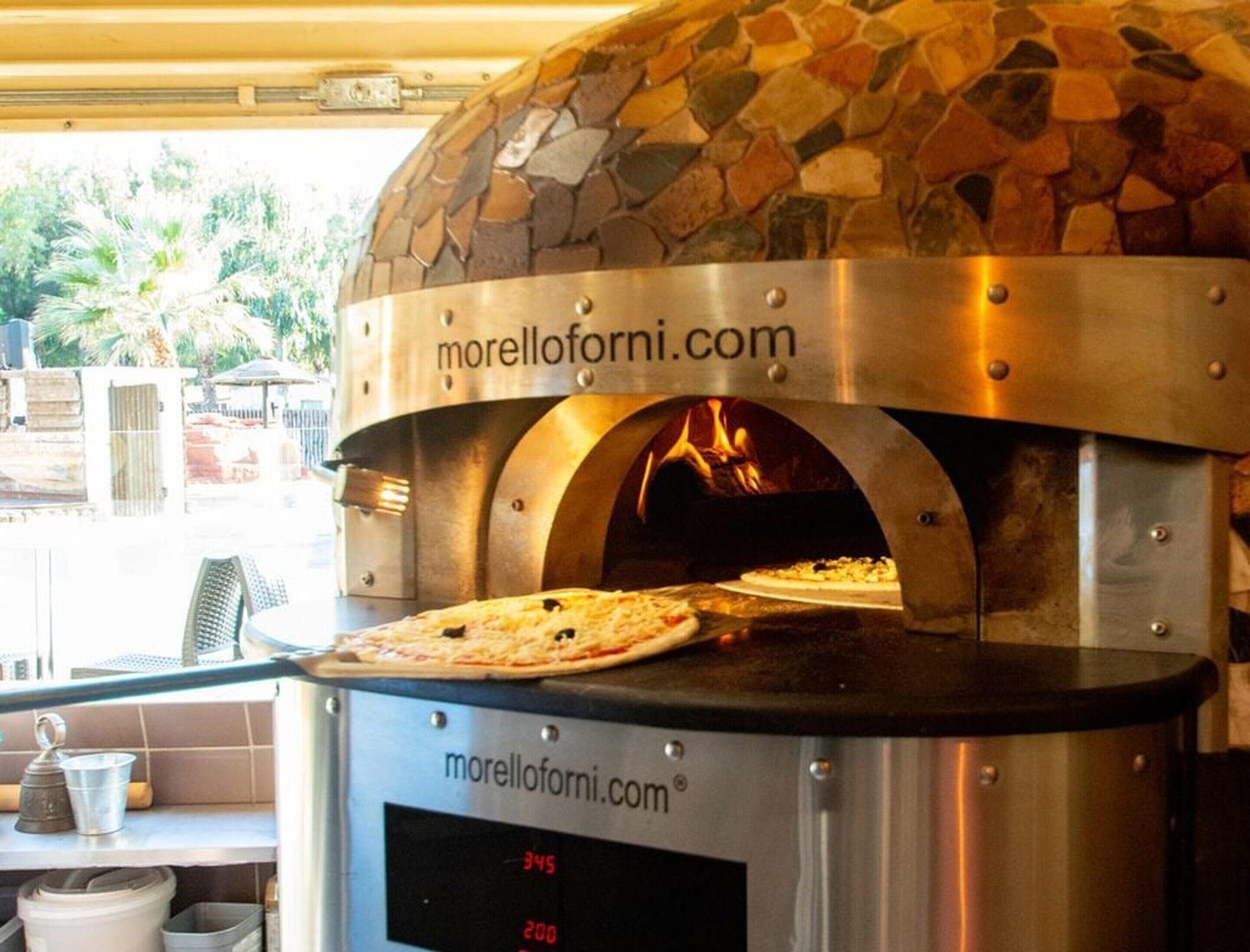 Horno para pizza al aire libre con baldosas de piedra, una pantalla digital, una pizza en su interior y un nombre de sitio web a la vista.