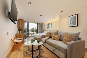 A modern hotel room with a sofa, a small table, a TV mounted on the wall, and a balcony with a view.