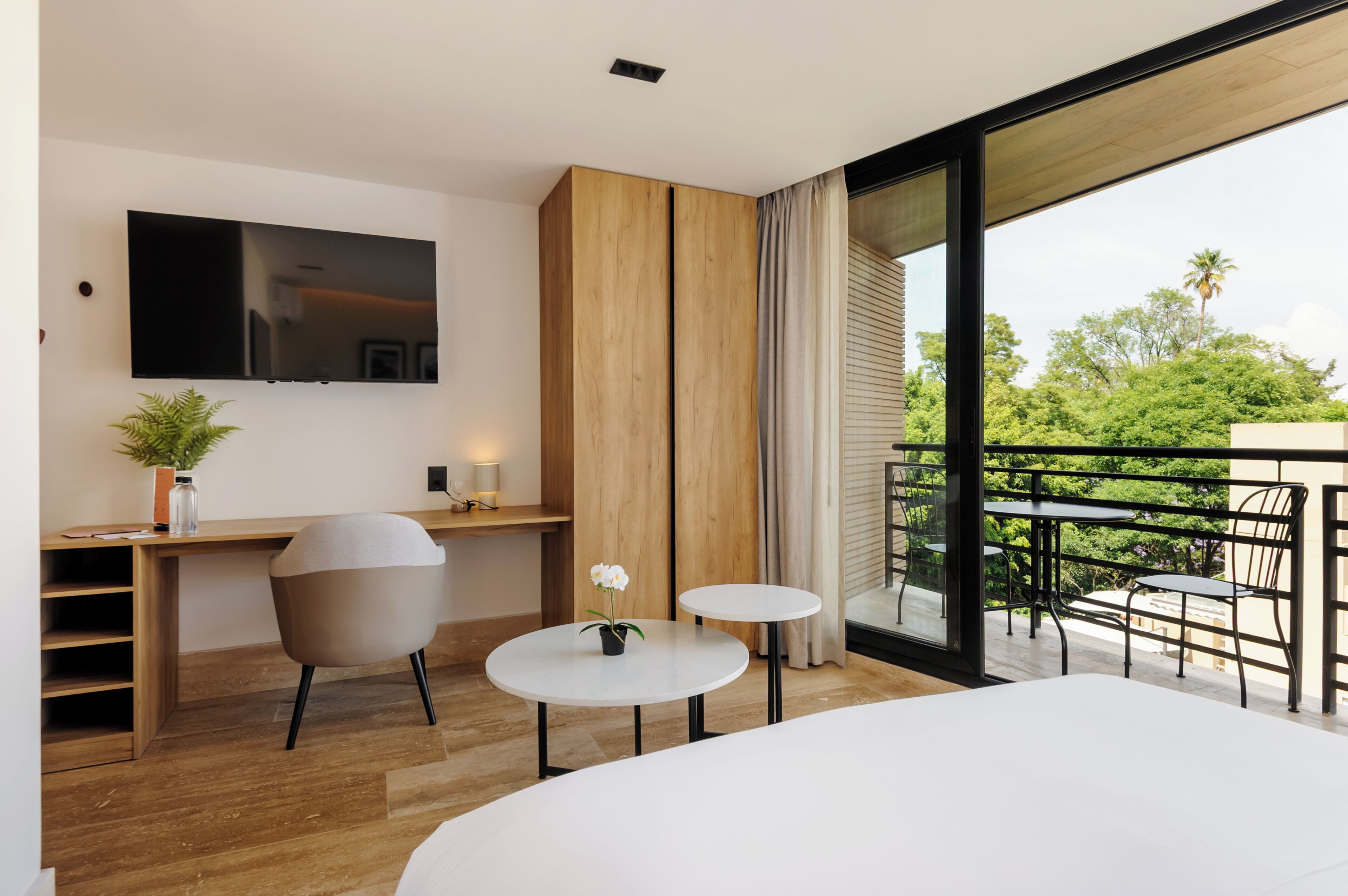 A modern hotel room with a wooden desk, a TV, a small round table with a vase, a chair, and a balcony with outdoor furniture.