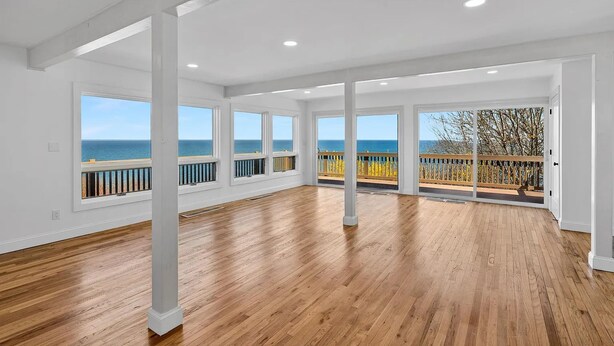 A spacious room with large windows, wooden flooring, and a balcony with a view of the sea. - Baiting Hollow Waterfront, Private Beach & Stunning Sunsets (Riverhead)