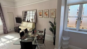 A modern dining area with a glass table, black chairs, a radiator, a window with blinds, and wall art. - Guildford Apartment with Free Parking & Garden (Guildford)