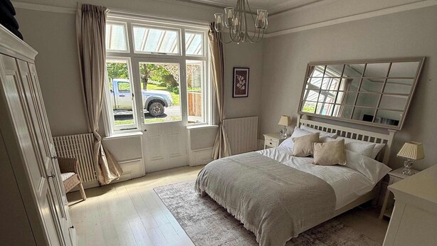 A bedroom with a bed, bedside tables, a chandelier, a window with curtains, a radiator, and a large mirror. - Guildford Apartment with Free Parking & Garden (Guildford)