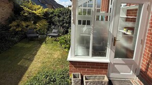 A conservatory with a glass door, a tiled patio, and a small garden with a lawn and a hedge. - Guildford Apartment with Free Parking & Garden (Guildford)