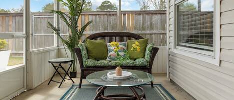 A covered patio with a glass table, wicker sofa with green cushions, a potted plant, and a view of the backyard.